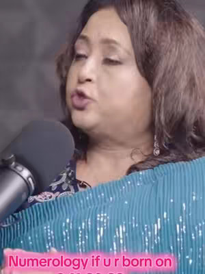 A woman with dark hair and light skin is speaking into a microphone. Text on the image reads 'Numerology if u r born on'. She is wearing a blue sequined top and her background appears to be a dark room. A microphone is present in the shot.