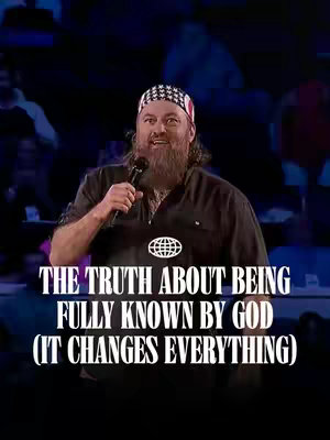 A man with a thick, dark beard and styled hair is holding a microphone, speaking in front of a blurred audience. He is wearing a black shirt with a complex pattern on the bandana he holds, and there's a simple logo that looks like the world in the center front of his shirt. Below him is an overlay of text over a dark, blurred background.