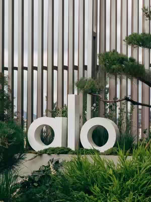 The image showcases the white 'alo' logo amidst lush greenery and shrubbery, set against a backdrop of vertical, alternating shades of brown and beige, creating a contrasting background. The logo, in a clean, modern font, appears to be a part of a sign, with the backdrop suggesting a building exterior. The scene gives the impression of a zen-like environment, with well-maintained plants and architectural elements.