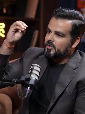 A man with dark hair and a trimmed beard speaks into a microphone, gesturing with his right hand. He wears a dark t-shirt under a gray blazer. The background suggests an indoor setting with a bookshelf and soft lighting.