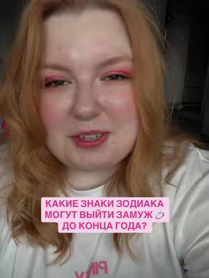A medium shot shows a woman with fair skin and reddish-blonde hair in a brightly lit setting. She's facing the camera, with a light expression, and wearing pink eyeshadow and blush. She is wearing a white shirt. A pink rectangular text box is positioned at the bottom, containing Cyrillic text. The background is out of focus, suggesting a simple indoor environment.