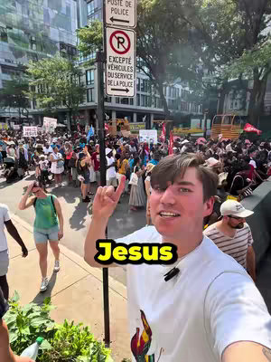 A selfie-style shot taken from a slightly low angle, featuring a young man against a backdrop of a vibrant outdoor crowd. He's smiling and pointing upward with one finger. The background shows a crowded street with multiple people, buildings, and signs in view, suggesting an urban setting. The main color theme seems to be a bright, sunny day with a mix of natural and urban elements.