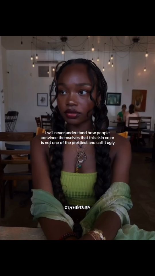A young Black woman is the focus, seated in front of a blurry background with low lighting. Her hair is styled in braids with curled ends, and she wears a neon-green top and a scarf draped over her shoulders. The text overlay features white text, highlighting a message about beauty and skin color. The background suggests a cafe or restaurant setting with light fixtures.