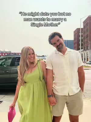 A couple stands close together, holding hands. The woman on the left wears a green dress and holds a pink clutch, while the man wears a white shirt and khaki shorts. Text overlayed at the top reads, "He might date you but no man wants to marry a Single Mother". The background includes a building and a car.
