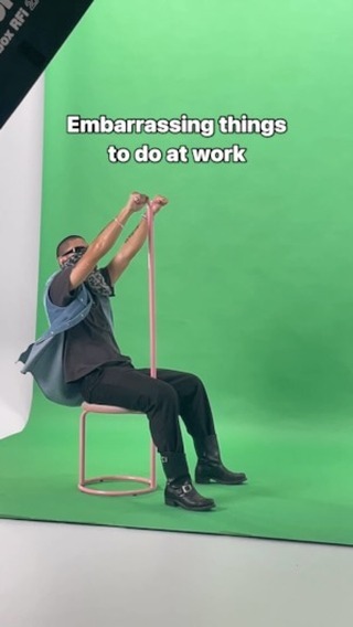 A person wearing sunglasses and a scarf over their nose and mouth sits on a pink chair in front of a green screen. They are holding a long pink object, likely a training band, above their head, with the ends held in both hands. The image has text overlayed across a green backdrop. A studio light is partially visible.