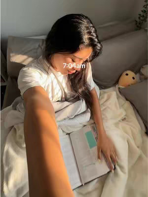 A young woman lies in bed, holding a book open. The bright morning sun streams in from the left, illuminating her and the room with a warm glow. The time, 7:04am, is displayed over her head. The setting is a peaceful bedroom with white bedding and gray pillows.