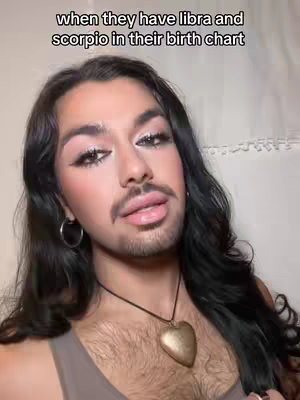 The image presents a close-up of a person with long dark hair, wearing makeup. The person has a beard and a pendant necklace. Text at the top reads 'when they have libra and scorpio in their birth chart.' The background is a light, neutral color.