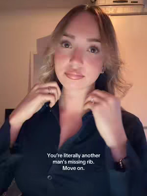 A woman in a dark blue button-down shirt looks towards the camera with a faint smile, her hands near the collar. The lighting is warm, with a text overlay at the bottom.
