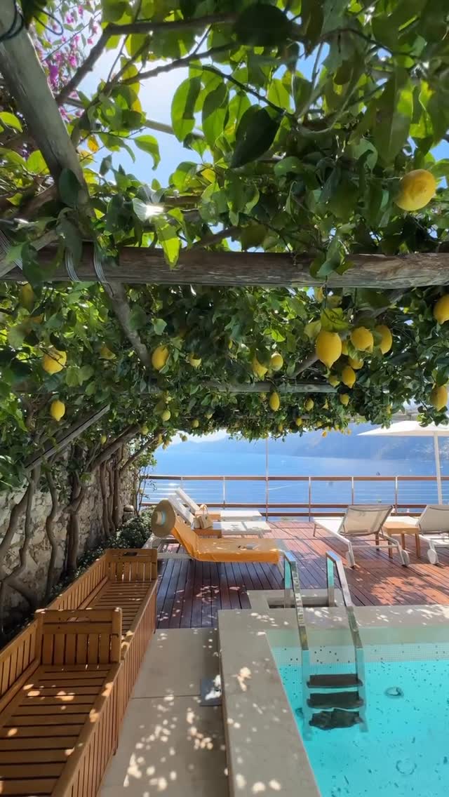 A scenic shot of a luxurious outdoor setting with a lemon pergola overhead. Yellow lemons hang from the branches, providing shade over wooden deck with lounge chairs and a small pool. The ocean is visible in the background.