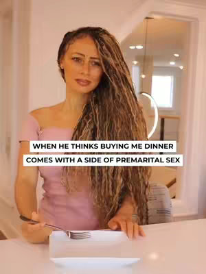 A woman with long, wavy hair and a pink dress is seated at a table, looking towards the camera. On the table in front of her is a white plate with a fork on it. Overlay text reads: "WHEN HE THINKS BUYING ME DINNER COMES WITH A SIDE OF PREMARITAL SEX". The background is blurred.