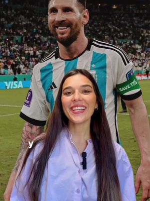 A woman smiling at the camera with a background image of soccer player Lionel Messi. The woman is wearing a light purple button down collared shirt, and the soccer player is wearing his soccer uniform. The background shows the crowd behind the soccer player.