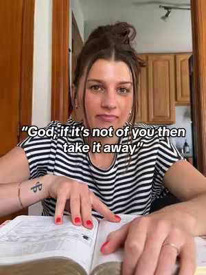 A medium shot shows a woman looking directly at the camera, hands resting on an open book. She has dark hair in a high bun and is wearing a striped shirt. The setting is a room with wooden trim and a cabinet visible in the background.