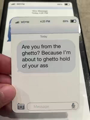 The image displays a close-up of a white card held in a hand, resembling a phone message interface. A second, less focused phone screen provides a blurry backdrop. The card has a text bubble displaying the phrase "Are you from the ghetto? Because I'm about to ghetto hold of your ass."