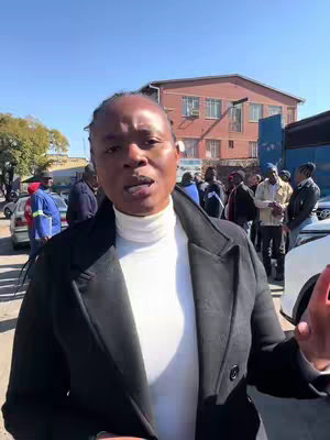 A woman wearing a white turtleneck and a black coat is speaking to the camera, likely addressing a crowd of people blurred in the background. The scene appears to be outdoors on a sunny day.