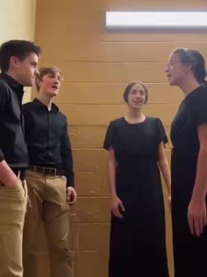 The image shows four people, two men and two women, standing in front of a light brown, paneled wall. They are arranged in a loose semicircle and appear to be singing. The lighting is simple and the scene is a medium shot. The people appear to be of mixed ages, and the color scheme consists of earthy tones and dark clothing.