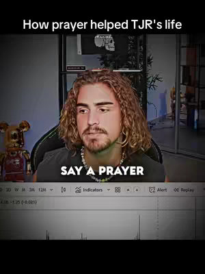 The cover image features a man in the center, looking downwards with a thoughtful expression. Text overlays read 'How prayer helped TJR's life' at the top and 'SAY A PRAYER' in bold white letters at the bottom. The man has curly brown hair, a short beard, and is wearing a dark shirt with a beaded necklace. Behind him, a room with a computer setup is vaguely visible. There is a trading chart at the bottom of the screen.