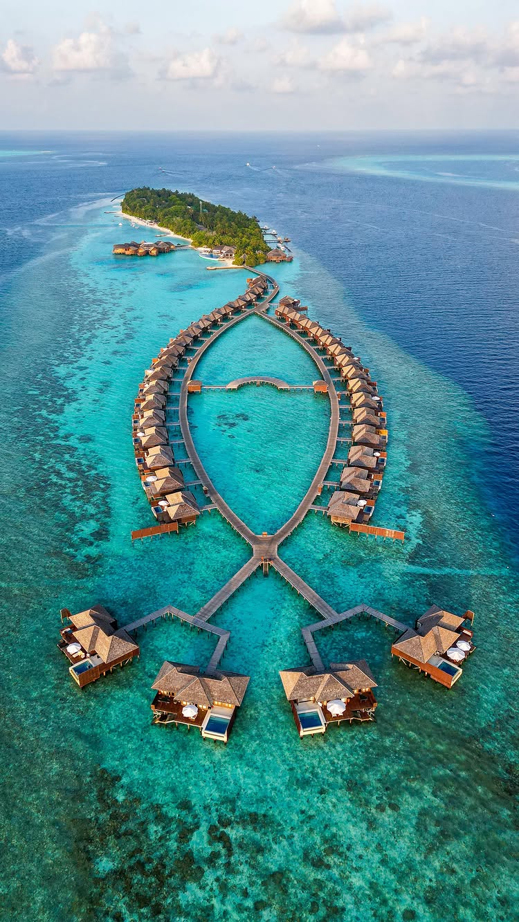 An aerial shot captures a stunning tropical scene. Overwater bungalows, each with a thatched roof, are arranged in a symmetrical pattern over crystal-clear turquoise water. They extend from walkways that converge into a complex wooden structure. A vibrant green island, dotted with lush vegetation and additional bungalows, provides a backdrop, and the deep blue ocean surrounds the entire scene.