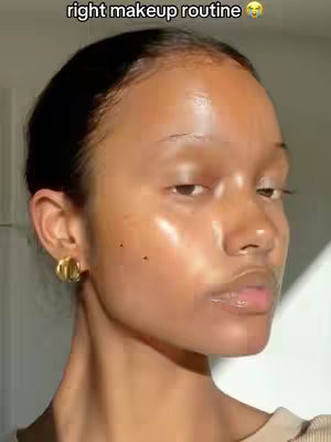 A close-up shot of a person's face, demonstrating a makeup routine. The individual is positioned slightly off-center, with their face illuminated to highlight the dewy makeup application. The light accentuates features like the lips and the overall glow of the skin. The background is simple and out of focus, placing all attention on the face.