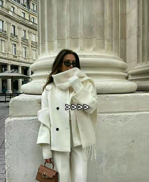 A woman wearing sunglasses and a white outfit, posing in front of a large white column. She is holding a small light brown purse. The background includes more architectural elements and a glimpse of greenery.