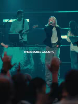 A live music performance scene with a blonde female singer in the center, wearing a black blazer and white shirt, holding a microphone. A band is behind her with a guitarist, and a crowd of people is in the foreground with their hands up. The background is dimly lit with blue and green hues, creating a stage environment.