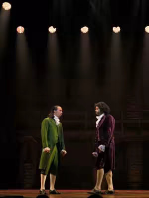 Two male figures stand facing each other on a stage. The stage is dimly lit, with spotlights overhead. Both figures are dressed in period clothing, suggesting the setting is a historical play or musical. The figure on the left is in a green coat and the figure on the right is in a burgundy coat.