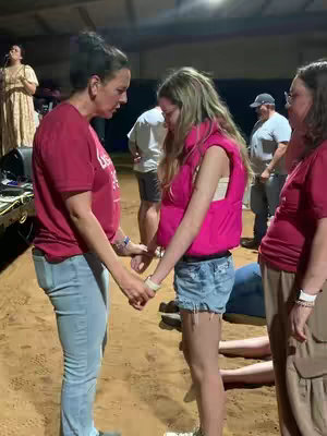 The image shows a group of people in a dimly lit indoor setting, likely a meeting or event. Two women are the central focus, holding hands as if in prayer or support. A person with long hair wearing a pink vest and denim shorts, with eyes closed, is being supported by the two women. Other people in the background also seem to be partaking in a group event. The lighting appears to be from overhead sources, casting shadows and highlighting the subjects.