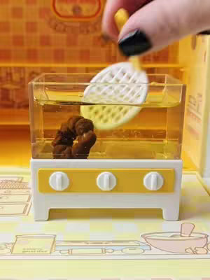 A close-up shot of a miniature kitchen setup with a clear container filled with a yellow liquid, likely oil. Inside the container, there is a small, dark donut partially submerged. A white, slotted spatula is being dipped into the oil next to a small, dark, cooked donut, as a hand is holding up a stick with a yellow object on its end. The background features a yellow and orange checkered pattern. The lighting appears bright and focused towards the center. The composition is visually appealing, with a focus on the miniature food preparation.
