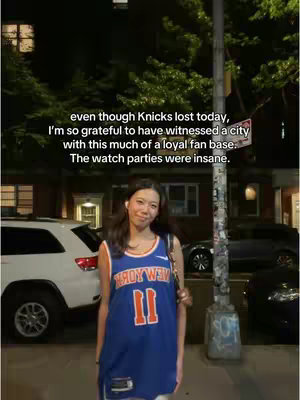 A young woman stands on a city street at night, wearing a New York Knicks jersey. Behind her are parked cars and buildings, with text overlaid that describes the city's support for the team.
