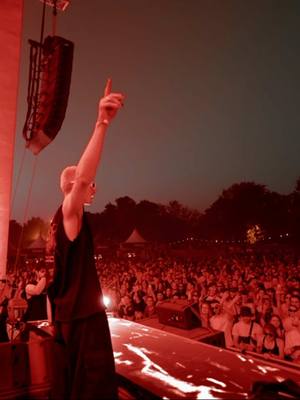 Image captures a concert or festival scene at night. A person, lit with red lights, is the central figure, positioned slightly off-center and facing away from the viewer. Their right arm is raised, index finger extended towards the sky. The background reveals a large crowd of people with a stage set. The overall tone is energetic, suggesting an exciting live event.