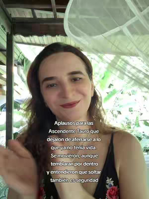 A selfie-style shot of a woman with long brown hair, angled upwards. The woman is smiling slightly with red lipstick and is slightly clapping her hands. The background shows nature.