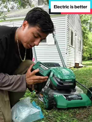 A man is kneeling on a lawn next to a green and black electric lawn mower. The background includes a white house and green grass. The man is holding a black battery and seems to be inserting it into the mower.