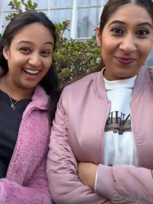 Two women are smiling in a selfie-style shot, positioned closely together. One on the left wears a black top under a pink fluffy jacket, while the other on the right wears a white t-shirt under a light pink bomber jacket. They are positioned in front of an out-of-focus background of a window and some green foliage.