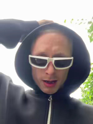 A person wearing a black hoodie and white-framed sunglasses looks directly at the camera against a background of foliage and a bright sky. The person has light skin and short hair, peering over the sunglasses with an open mouth.