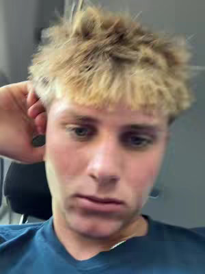 A close-up shot of a young man with light-colored hair. He is wearing a blue shirt and has a somewhat neutral expression. His hand is placed near his hair, and parts of a dark surface (likely a chair) are visible in the background. The lighting appears to be soft.