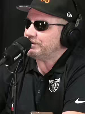 A man wearing a black baseball cap, sunglasses, and headphones is speaking into a microphone. He is wearing a black polo shirt with a logo on the chest.