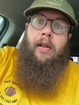 A close-up shot of a man inside a vehicle. He is wearing a green baseball cap and glasses. He has a thick beard and is wearing a yellow shirt with a football logo. The background shows parts of the car's interior, like the window and the door frame.