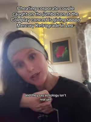 A woman with dark hair and a light blue headband is filming herself indoors. She's looking towards the camera and talking. Text overlays on the image include phrases about astrology and Mercury retrograde, and a framed picture with a floral design hangs in the background. The scene is lit with warm lighting and appears to be set in a home.