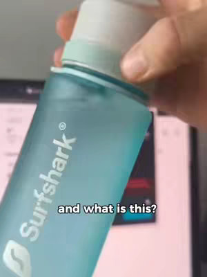 The video shows a close-up of a light blue water bottle being held in the frame. The bottle has the "Surfshark" logo on it. The text overlay reads "and what is this?" The background is blurred but seems to include a laptop screen.