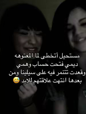 The image is a blurry, dark photograph featuring two women. The foreground is slightly out of focus, with the face of a woman on the left partly visible, showing a pained expression. The other woman's face in the right is partially visible and is smiling. Overlaid on the image is Arabic text.