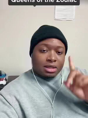 A man in a black beanie and a greyish sweater is looking at the camera and pointing upwards with his right index finger. There are white wired earbuds and a plain background.