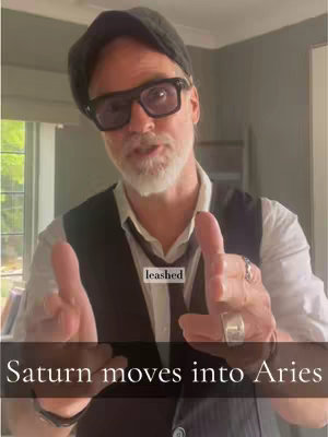 A man with a beard, glasses, and a hat points towards the viewer. Text overlays read 'Saturn moves into Aries.'