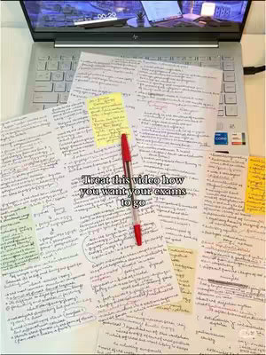 A top-down view of a workspace with a laptop, notes, and a pen. The laptop is open, displaying what appears to be a dark screen. Scattered across the table are multiple sheets of handwritten notes, with yellow sticky notes and a red pen lying on top. The composition has a slightly tilted angle, and is brightly lit.