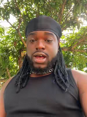 A man with locs and a black cap is the central focus, looking towards the camera with an open mouth, set against a blurred background of green foliage and branches.