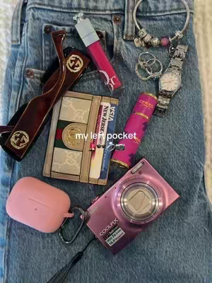 A close-up, slightly angled view of various items displayed casually on a denim fabric. In the center lies a wallet with a beige and green design, appearing to hold cards. To the left rest a pair of brown, Gucci-branded sunglasses and a pink AirPod case with its lid open. Adjacent to the case is a pink digital camera, while a pink lipstick tube rests beside a compact perfume. A silver bracelet and a silver watch are also present, adding to the ensemble. The text overlayed reads: 'my left pocket'. The color scheme is a mix of pinks and browns against the blue denim.