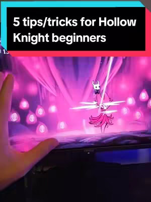 The cover image shows a screen displaying the Hollow Knight game. A text overlay at the top says '5 tips/tricks for Hollow Knight beginners' on a black background with a cyan outline. A hand is extending to the left, seemingly interacting with the display. The game screen has a predominantly pink and purple color scheme with a character from the game in the center .