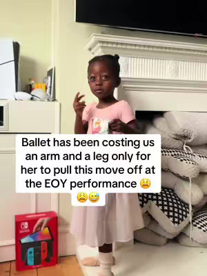 A young girl in a pink dress and white socks is standing indoors, seemingly in a living room due to the large ornate fireplace in the background. She is looking towards the camera, raising her finger to the side as if she's making a statement. A Nintendo Switch box can be seen on the floor to the left, indicating a gift or product placement. The image has a bright feel with off-white walls and a light-colored floor, with the girl as the central, most vibrant element.