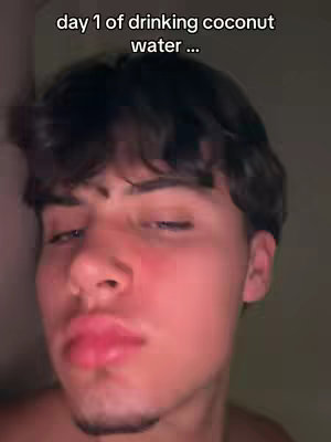 The cover image features a close-up of a young man's face, lit with warm tones that suggest a soft light source. He has short, dark hair and appears to be looking downward with a slight, thoughtful expression. His lips are full and a bit parted, contributing to a slightly pensive or even surprised look. The background is out of focus, suggesting a casual setting or low-light environment. The text overlay indicates reference to drinking coconut water.