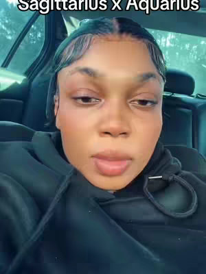 A selfie of a woman seated inside a car, framed from the chest up. She has dark skin, black hair with delicate baby hairs styled around her forehead. She looks slightly downward with a neutral expression, lips slightly parted. She is wearing a black hoodie. The background shows the inside of a car, with a window and a seat with a headrest.