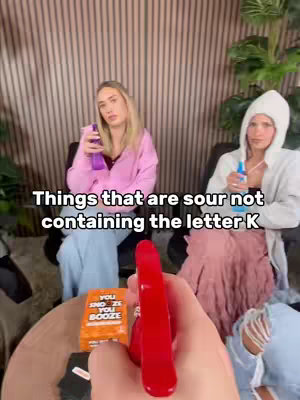 A first-person perspective shows a hand holding a red spray bottle, with two women in the background, each holding similar bottles. The left woman wears a pink sweatshirt and the right woman wears a white hoodie. Text is overlayed on the image.