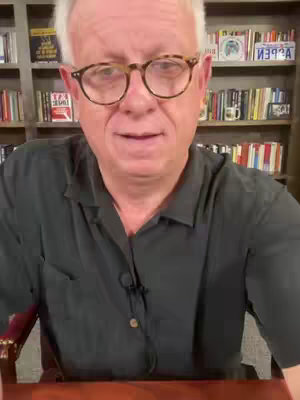 A man with glasses is centered in the frame, looking towards the viewer. He wears a dark green button-down shirt and a microphone is clipped to it. A bookshelf filled with books forms the background. The lighting appears to be natural.